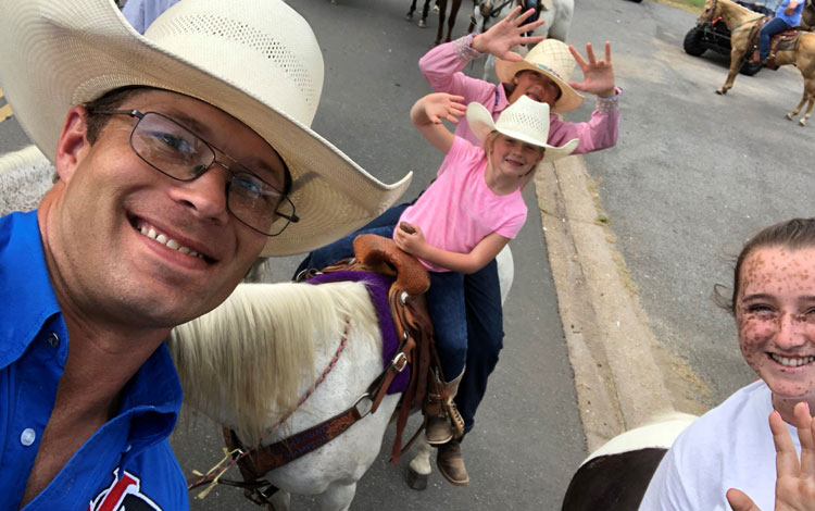 A cowboy taking a picture with two girls on a pony and another girl smiling.